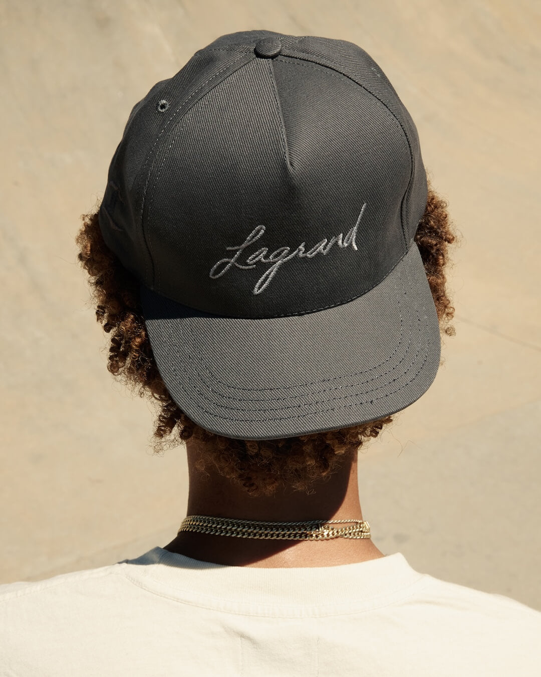 Model wearing the LAGRAND Script Hat in Cinder Grey, showing the front embroidered logo