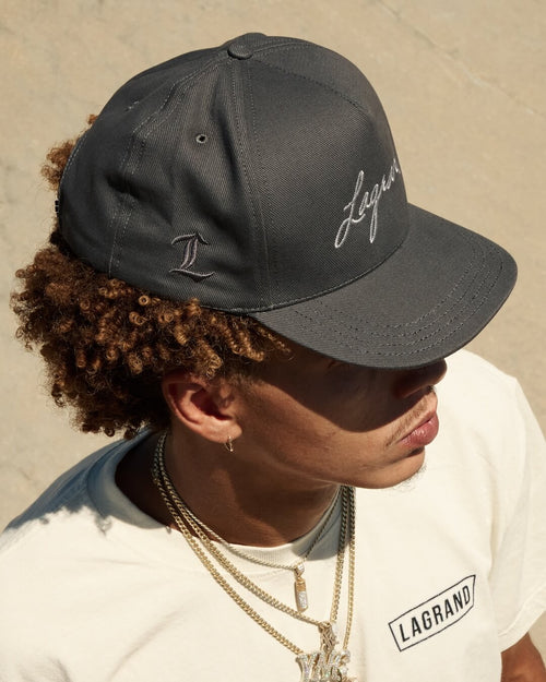 Model wearing the LAGRAND Script Hat in Cinder Grey, showing the side L embroidery